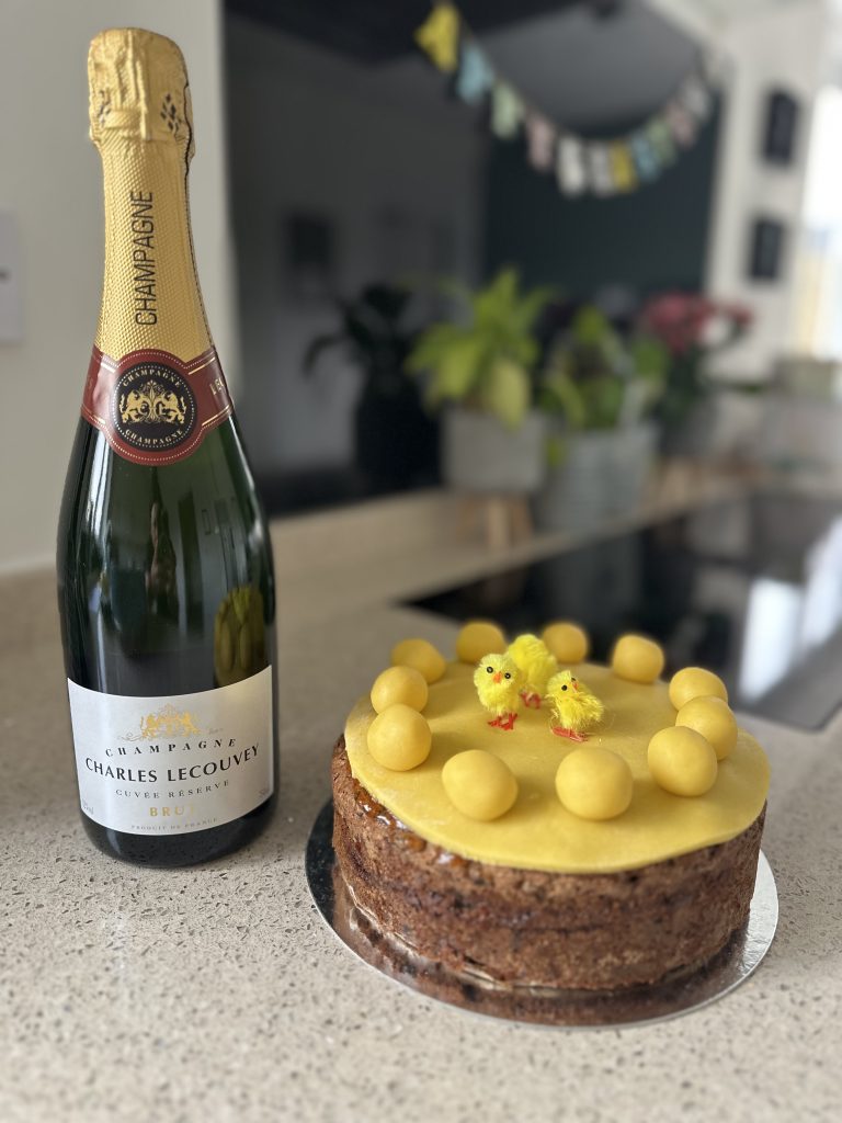 Bottle of champagne next to a homemade Easter fruit cake, topped with 11 marzipan balls and decorated with tiny yellow chicks – a celebratory treat from a family kitchen table. Thanks to Edmondsons IT for Sophie Seddon PR winning the champagne at the MYCC Business Festival last week.
