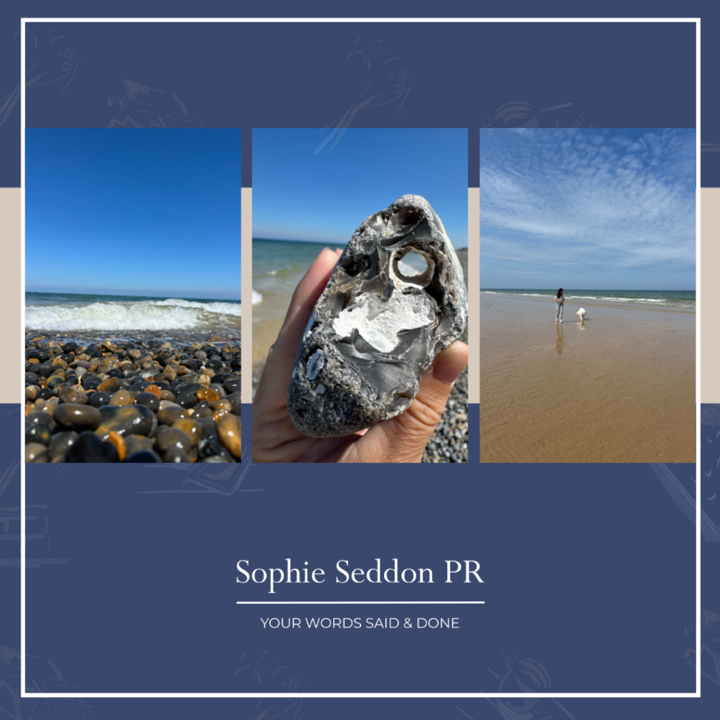 Pebble beach with sea tide washing in. Sophie’s hand holds up a rock with a natural hole, framing a different view. In the distance, her daughter and dog walk along the shoreline. Sophie Seddon PR logo appears at the bottom.